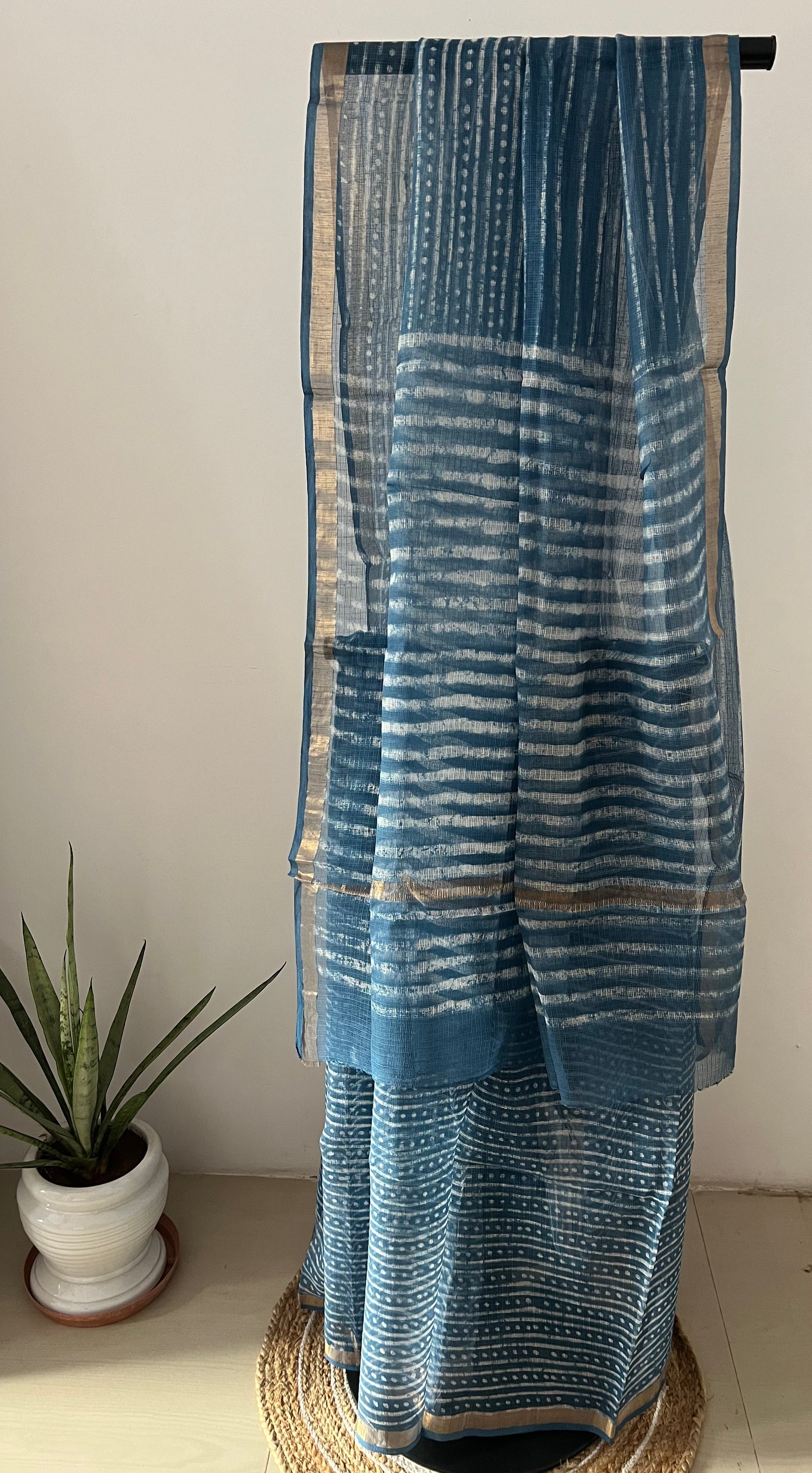 A blue patterned fabric, likely a saree or dupatta, draped over a stand in an indoor setting with a white wall and a small potted plant visible.